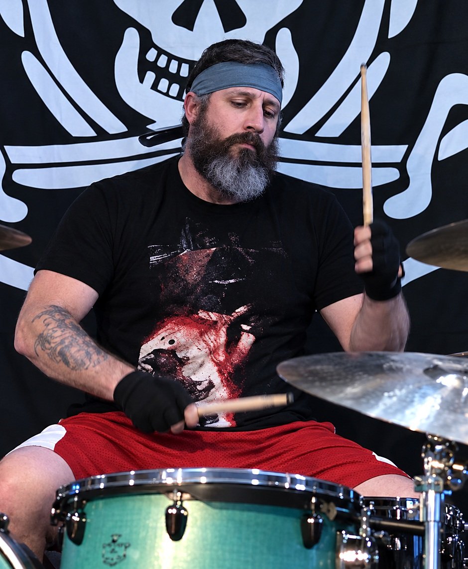 Brandon Aller, bearded and wearing a headband, playing drums on stage in front of a skull-and-crossbones backdrop.